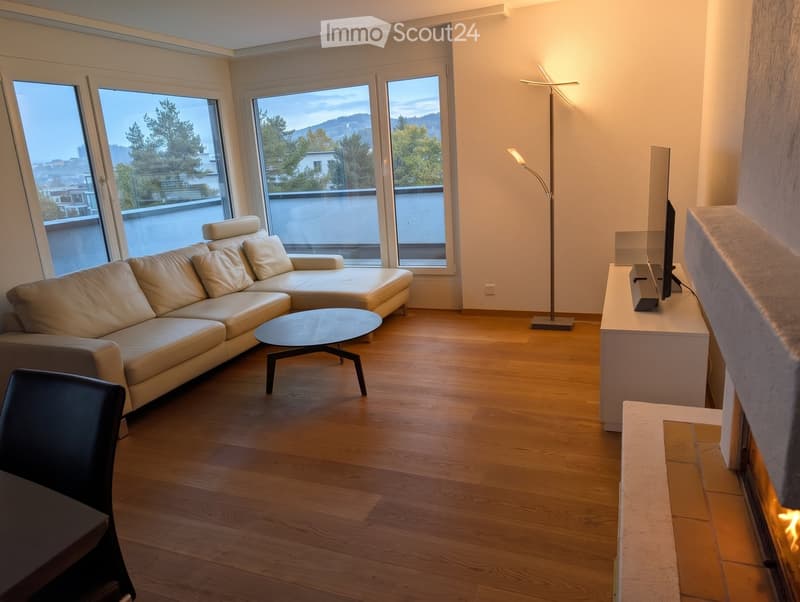 Modern living room with light wood flooring, large windows offering a scenic view, a beige sectional sofa, a round coffee table, a sleek floor lamp, and a fireplace with a TV on a white console.