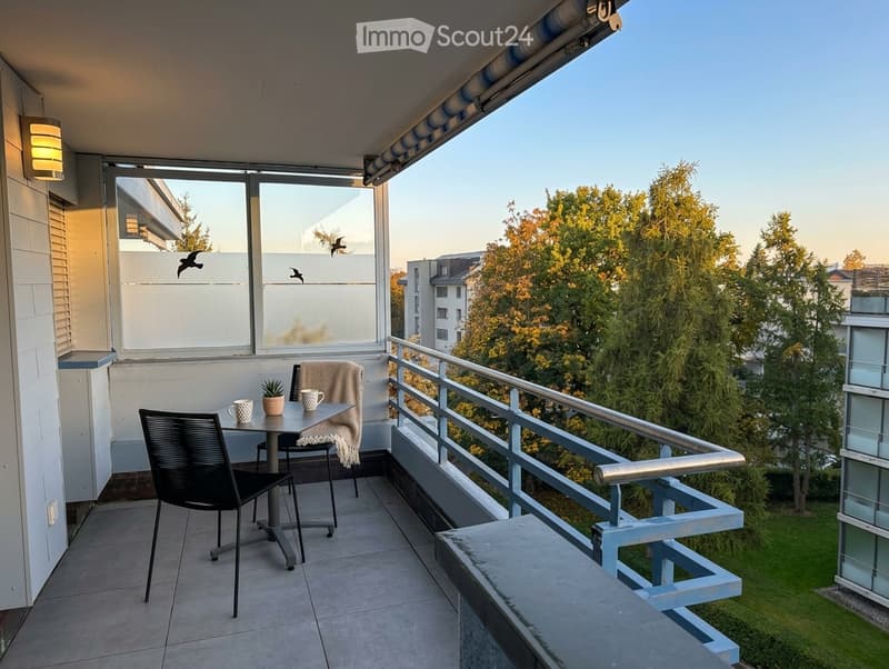Balcony with a small table, two chairs, a cup, and a small plant, overlooking a green area with trees.