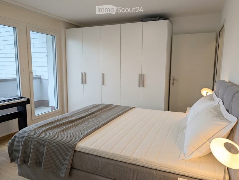 Modern bedroom with a large bed, white wardrobe, bedside lamp, and a small piano near the window.
