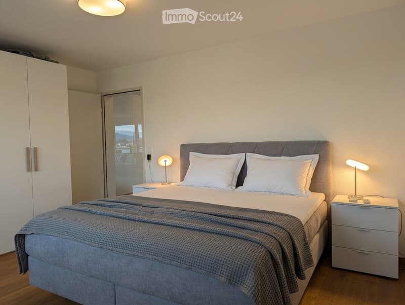 Modern bedroom with a neatly made bed, two nightstands with lamps, and a large wardrobe. The room features light-colored walls and wooden flooring.