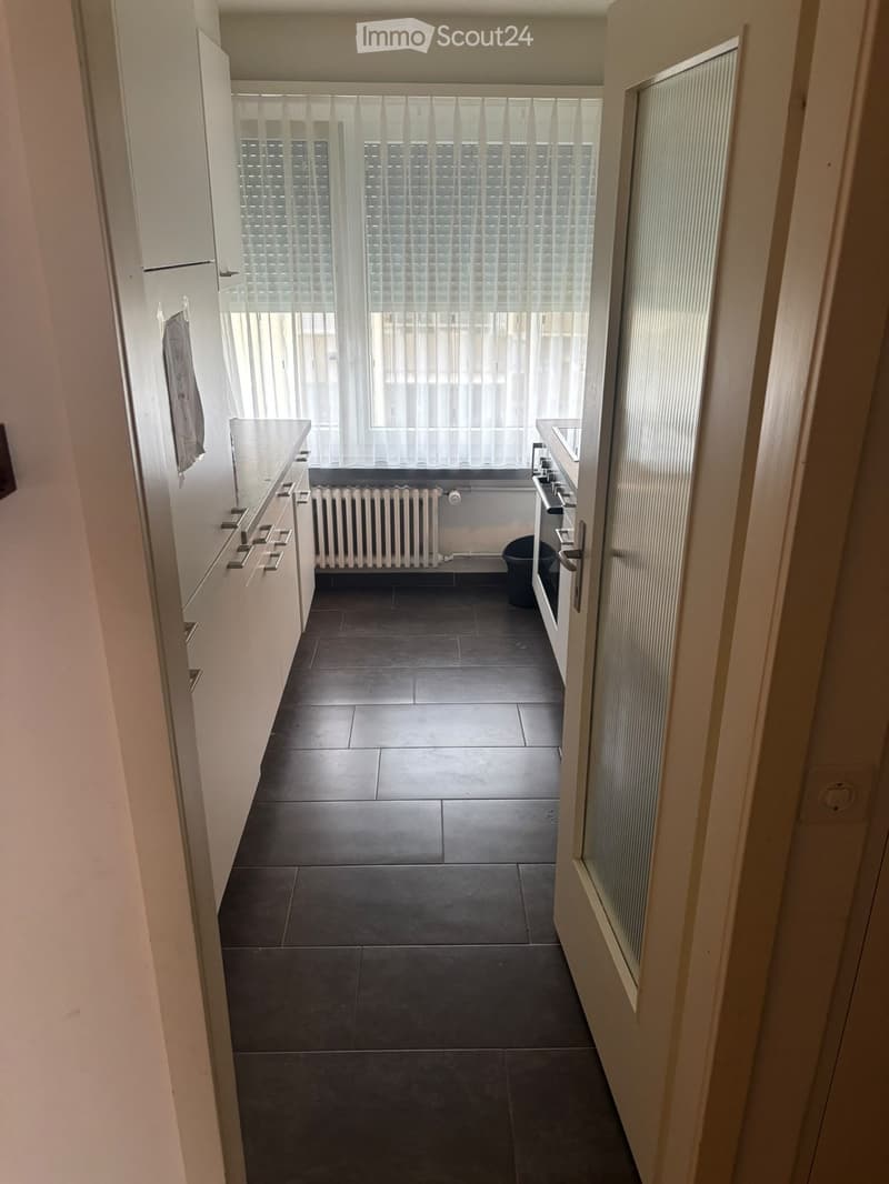 Empty kitchen with dark tiled floor, white cabinets, and a window with white blinds. No furniture or appliances are present.