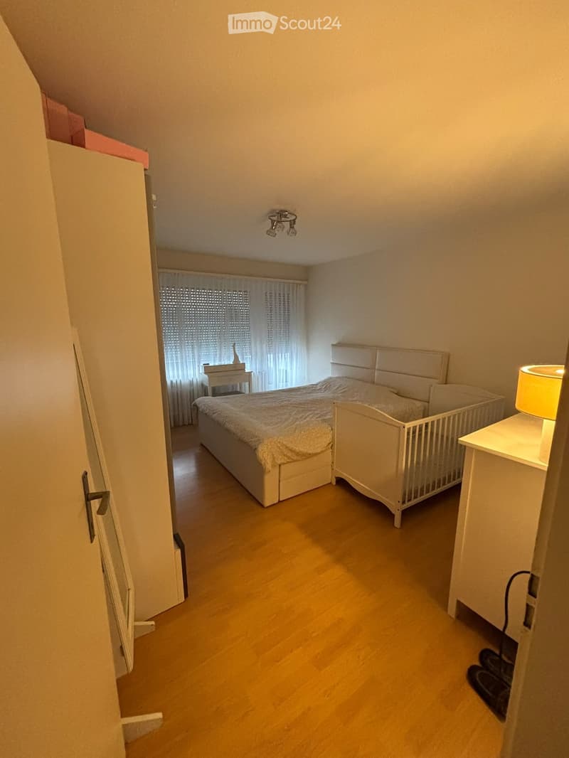 Cozy bedroom with wooden flooring, a large bed with white headboard, a nightstand with a lamp, a small table with a chair, and a crib in the corner. The room features a window with white curtains and a chandelier on the ceiling.