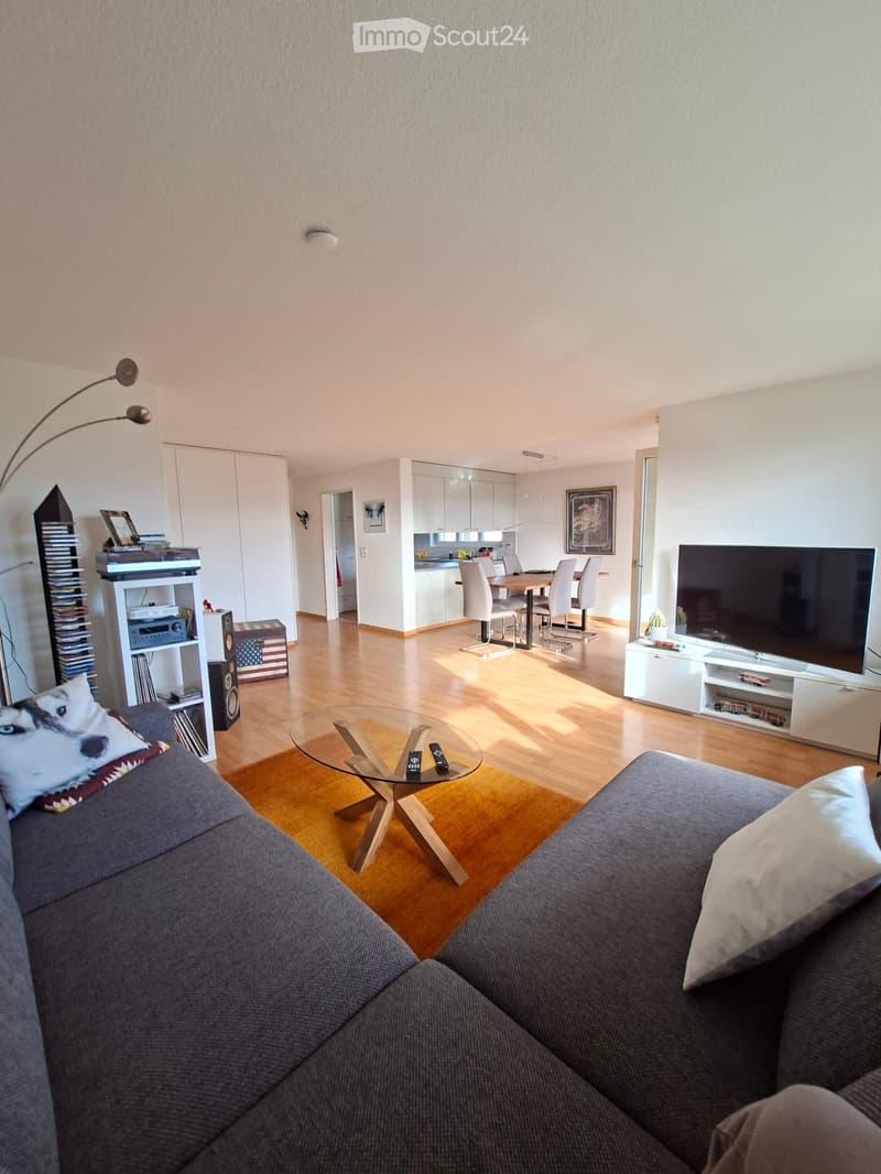 Modern living room with a grey sofa, glass coffee table, flat-screen TV, bookshelf, dining area with table and chairs, and a floor lamp.