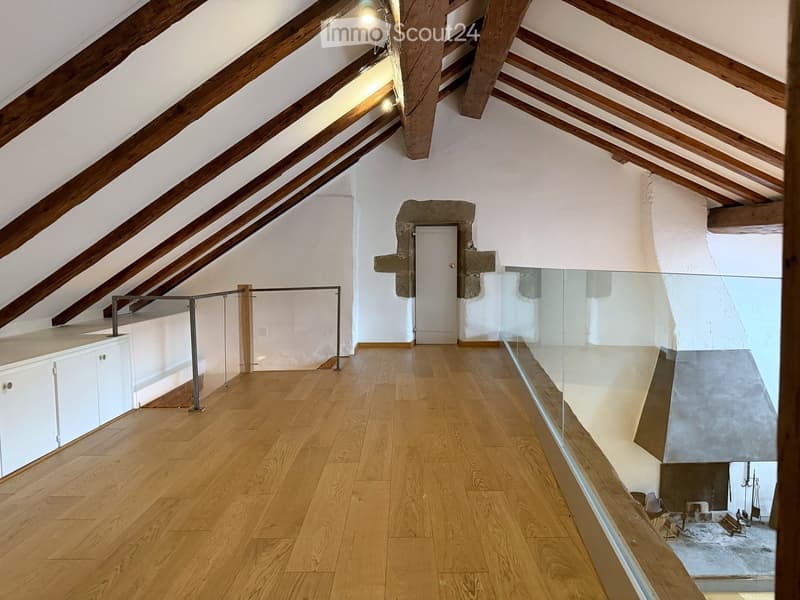 An attic space with wooden beams, wooden flooring, and a glass railing leading to a lower level. The room is empty with no furniture or furnishings.