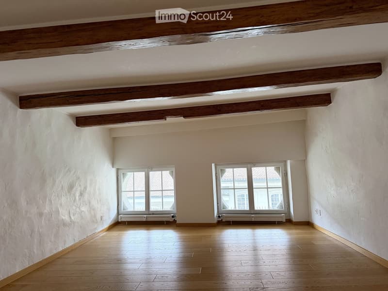 Empty room with wooden floor, white walls, exposed wooden beams on the ceiling, and two windows with white frames and radiators below them.