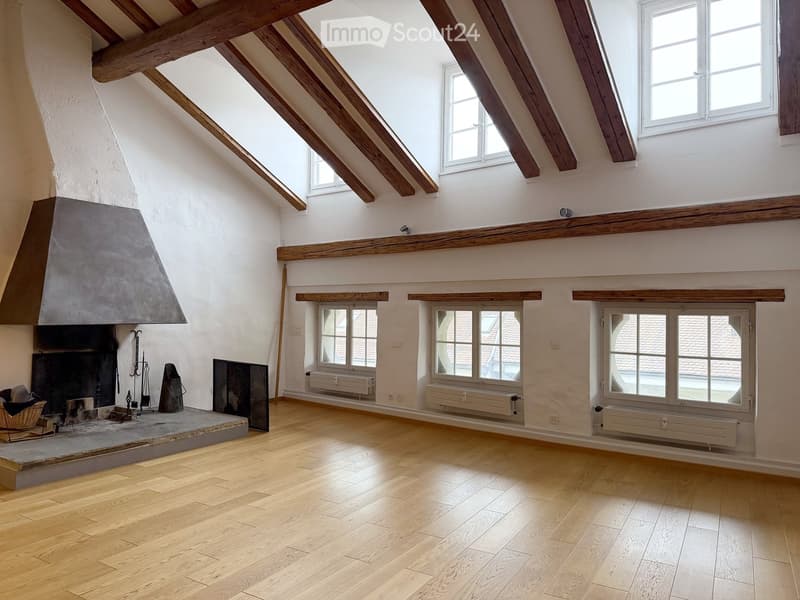 Empty attic room with wooden beams, exposed wooden floor, fireplace, and multiple windows.