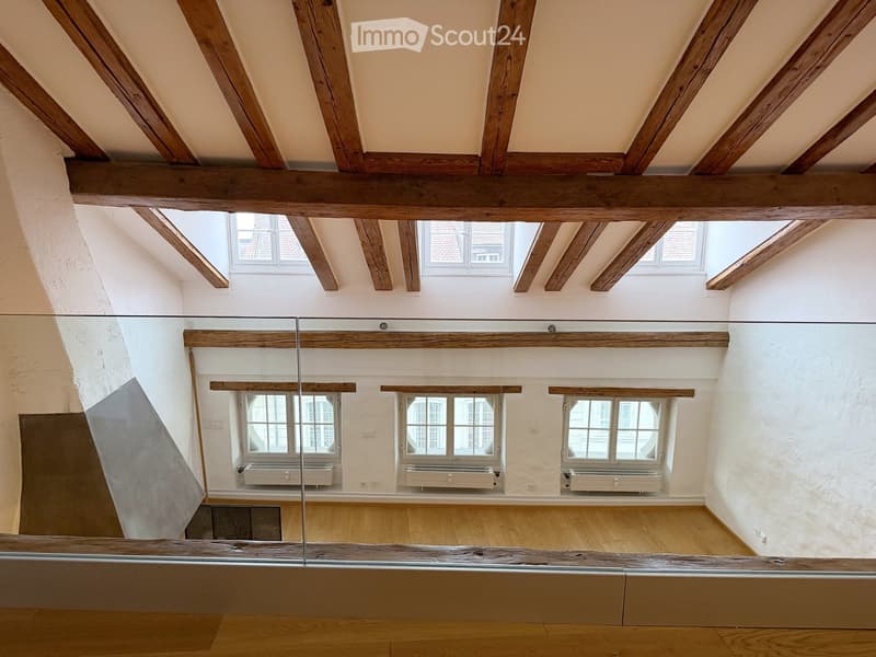 An empty room with exposed wooden beams, white walls, wooden flooring, and multiple windows, including skylights, with radiators beneath the windows.