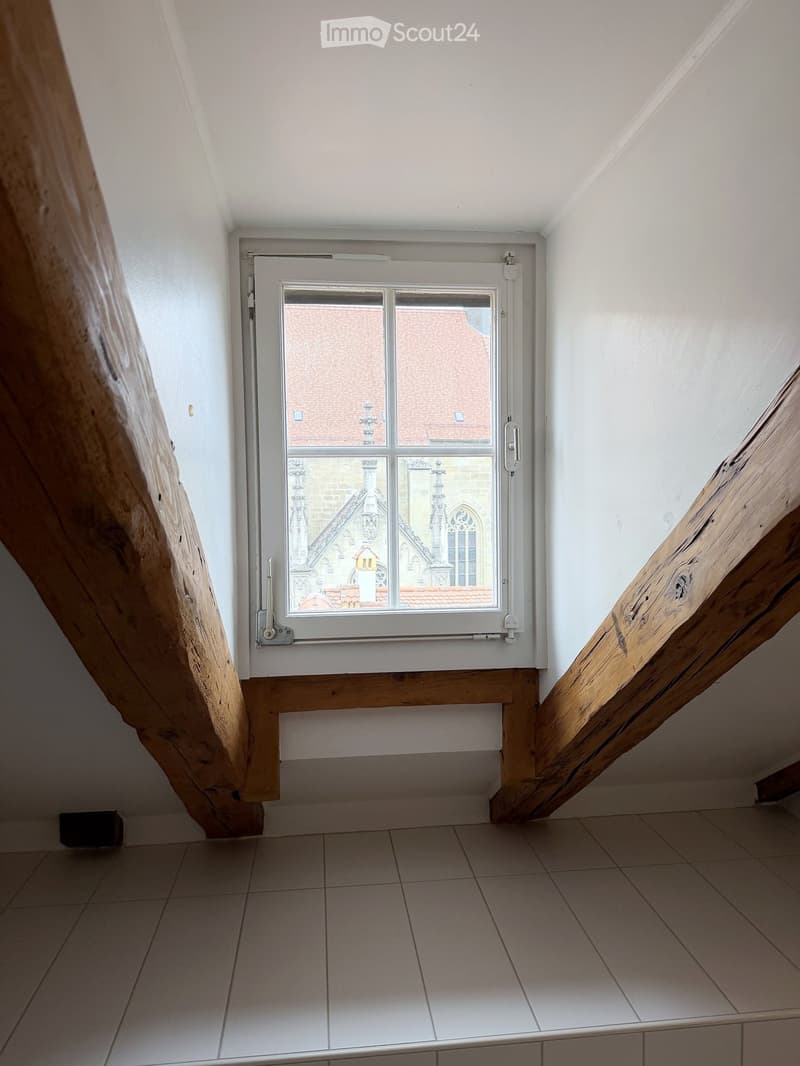 Empty room with tiled floor, white walls, and a window showing a view of a building with a red roof and architectural details.