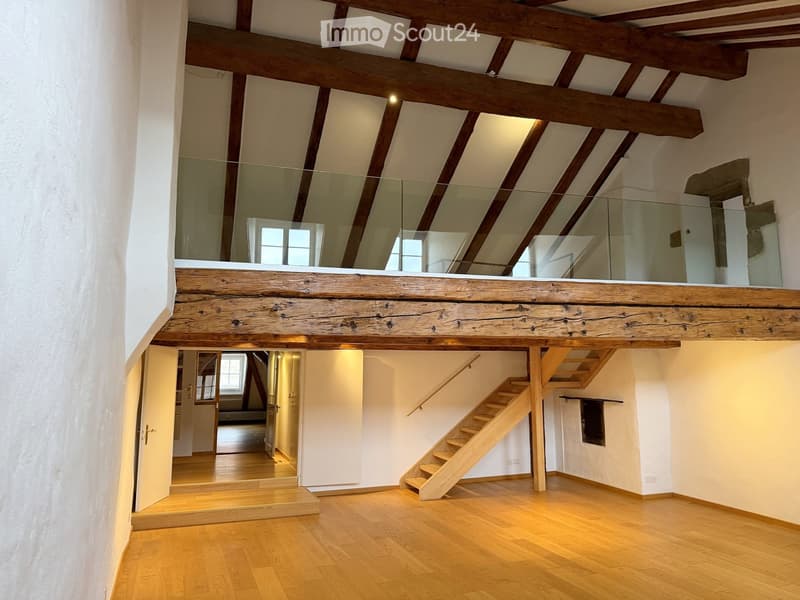 Open, airy room with wooden floors, exposed wooden beams, a staircase leading to an upper level, and a glass railing overlooking the space.