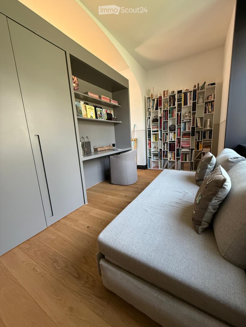 A modern, multi-functional room with a grey sofa, built-in bookshelf, desk, and a large cabinet. The room features wooden flooring and a slanted ceiling typical of an attic space.