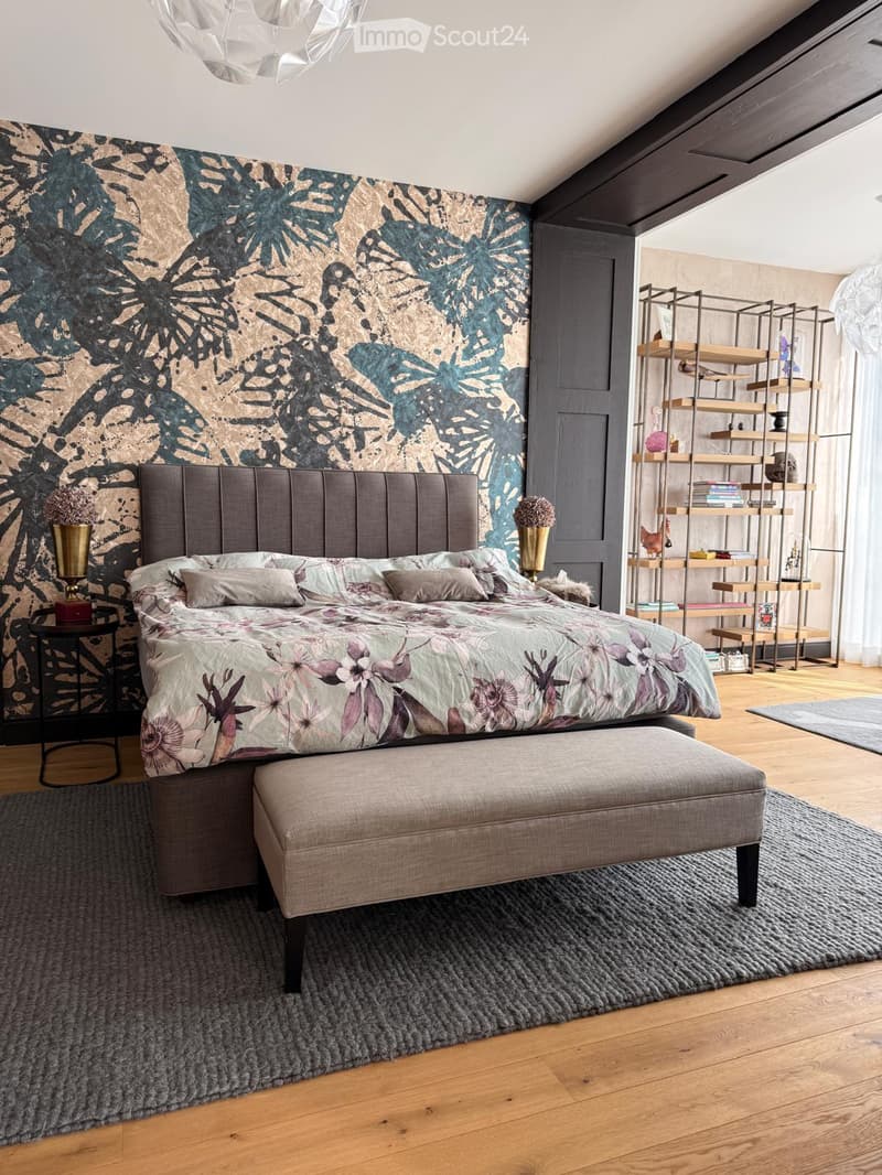 Modern bedroom with a large bed, floral duvet, nightstand, ottoman, and a decorative shelf unit. The room features a leafy patterned wall and wooden flooring.