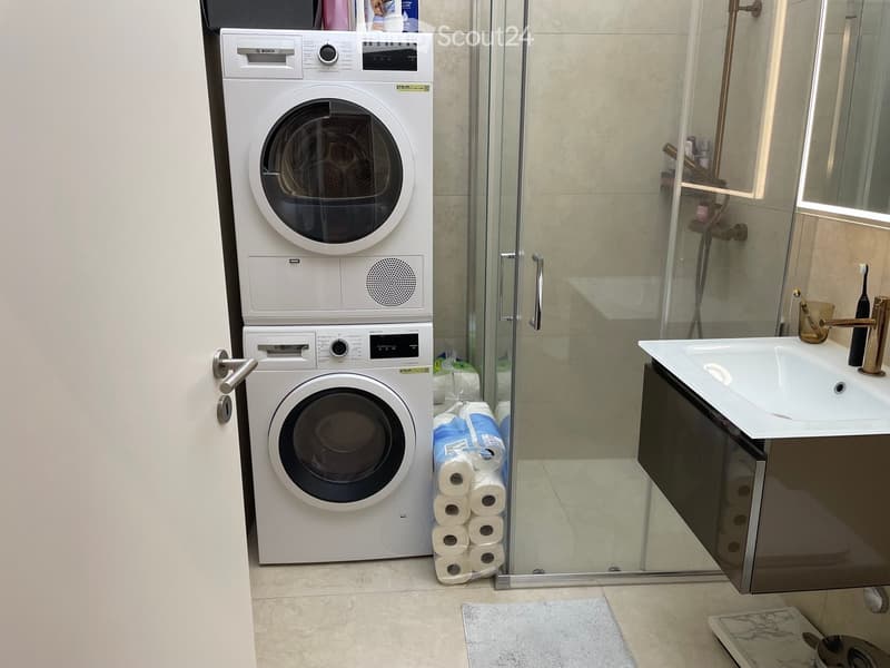 Modern bathroom with a washing machine and dryer unit, a sink with a modern faucet, a glass-enclosed shower, and a stack of toilet paper rolls on the floor.