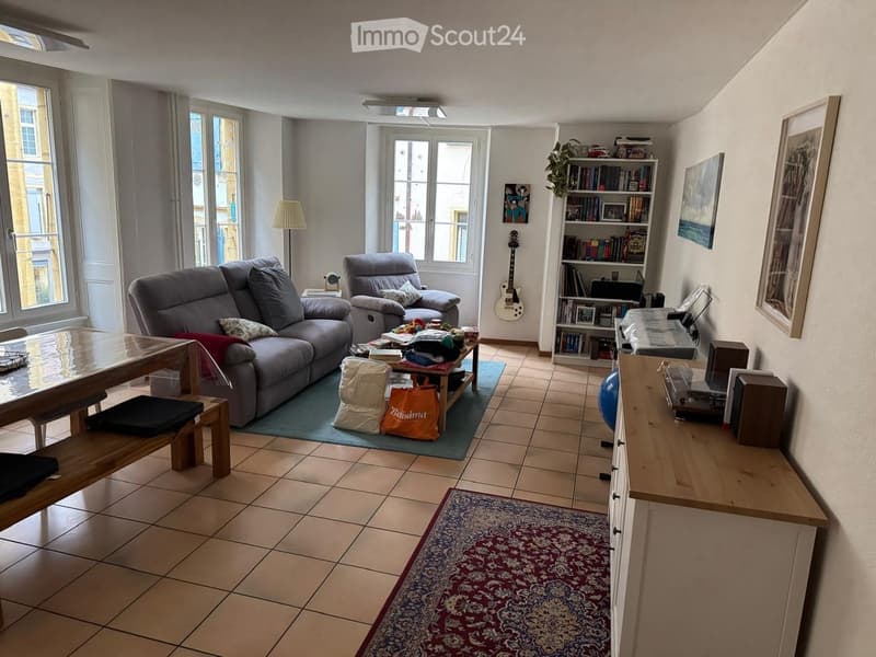 A modern living room with a grey sofa, armchair, coffee table, bookshelf, and a guitar on the wall. The room features large windows, tiled flooring, and a mix of decorative items including a lamp and a screen.
