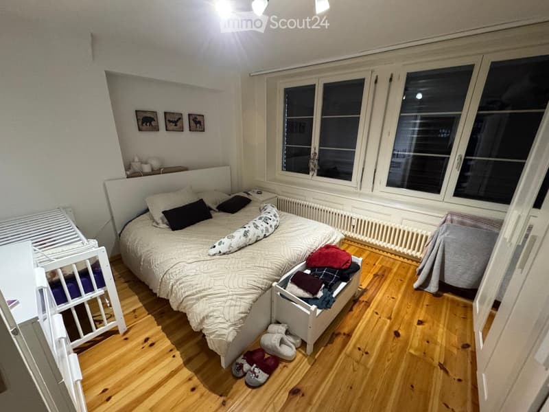 Cozy bedroom with wooden flooring, large window, bed with pillows and blanket, nightstand, radiator, picture frames, and a small stool with folded clothes.