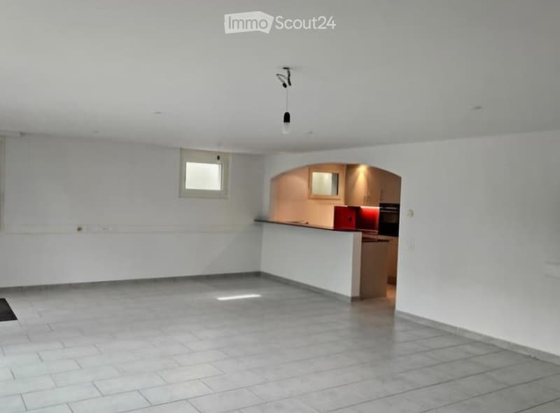 Empty room with white walls, tiled floor, and a small window. A doorway leads to a kitchen area with cabinets and appliances.