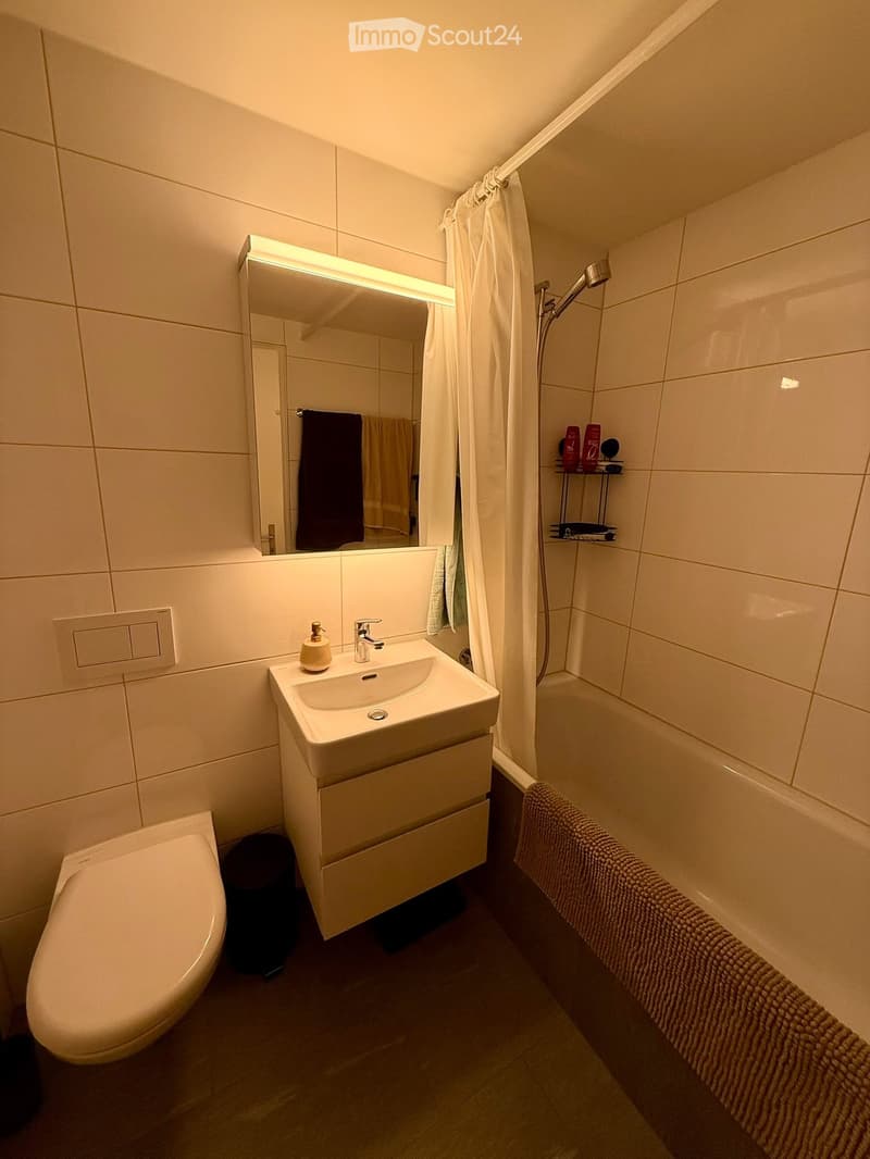 Modern bathroom with white tiles, a sink with a mirror, a toilet, a bathtub with a towel, and a showerhead.