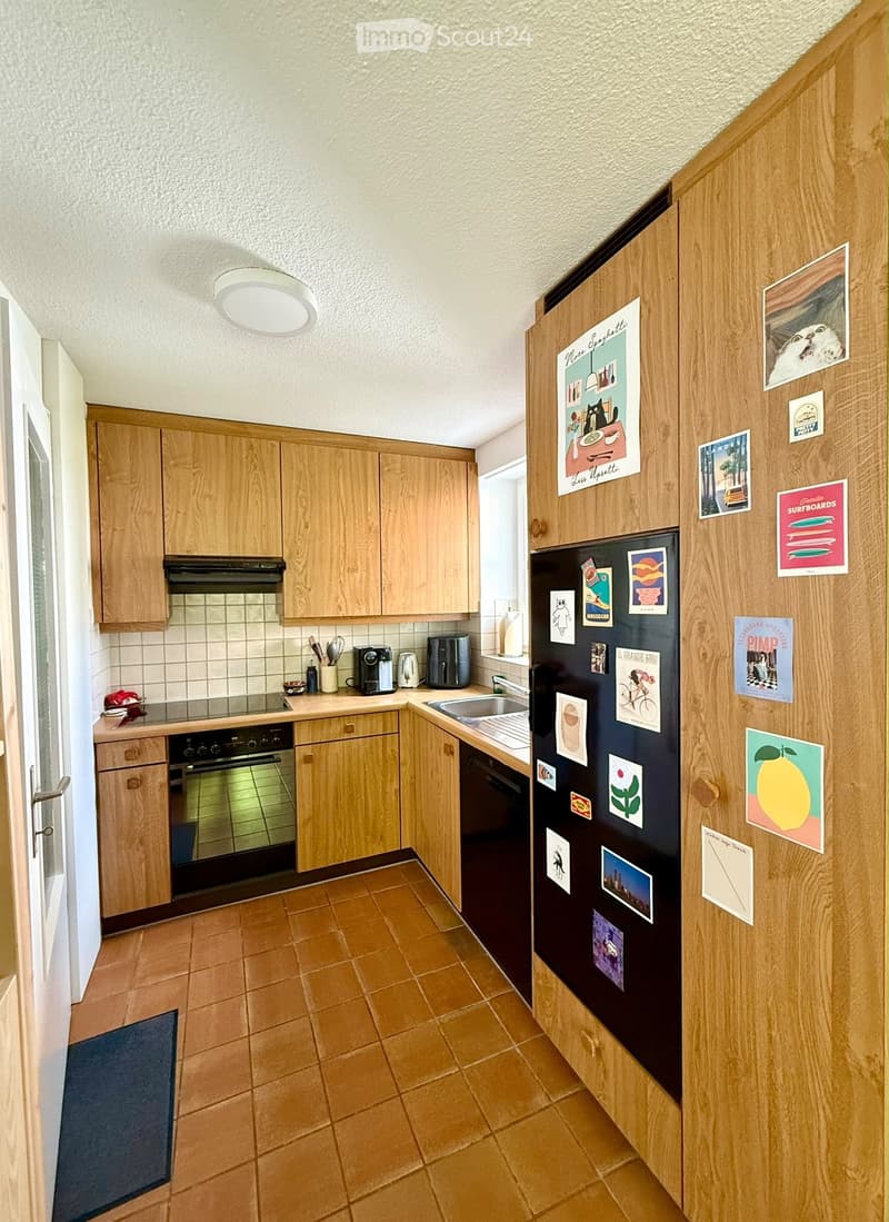 A cozy kitchen with wooden cabinets, a refrigerator covered in magnets and pictures, a sink, and various appliances including a coffee maker and a toaster. The floor is tiled in a warm brown color.