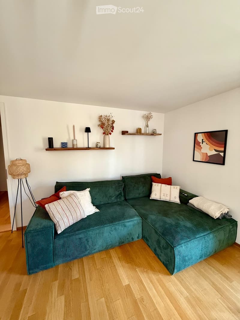 A modern living room with a green sectional sofa, wooden floor, decorative pillows, a floor lamp, and shelves with vases and artwork.
