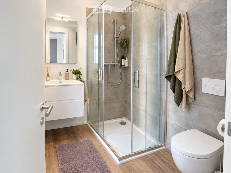 Modern bathroom with a glass-enclosed shower, white sink with mirror, toilet, and towels hanging on the wall. The space features wooden flooring and a sleek, contemporary design.
