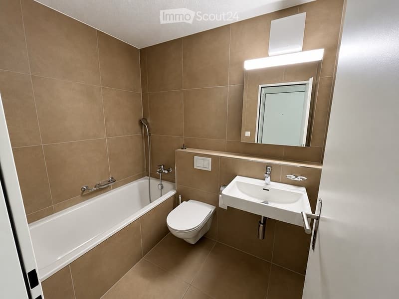 Modern bathroom with beige tiles, a bathtub, toilet, sink, and a large mirror with built-in lighting.