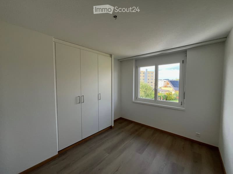 Empty room with light-colored walls, wooden flooring, and a large window with white frame. A white closet with double doors is visible on the left side.