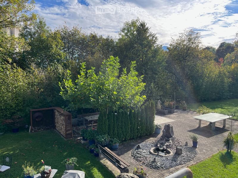 Well-maintained garden with a mix of lawn, paved pathways, and various plants. Features include a wooden bench, a small fountain, a table, and a storage shed for firewood.