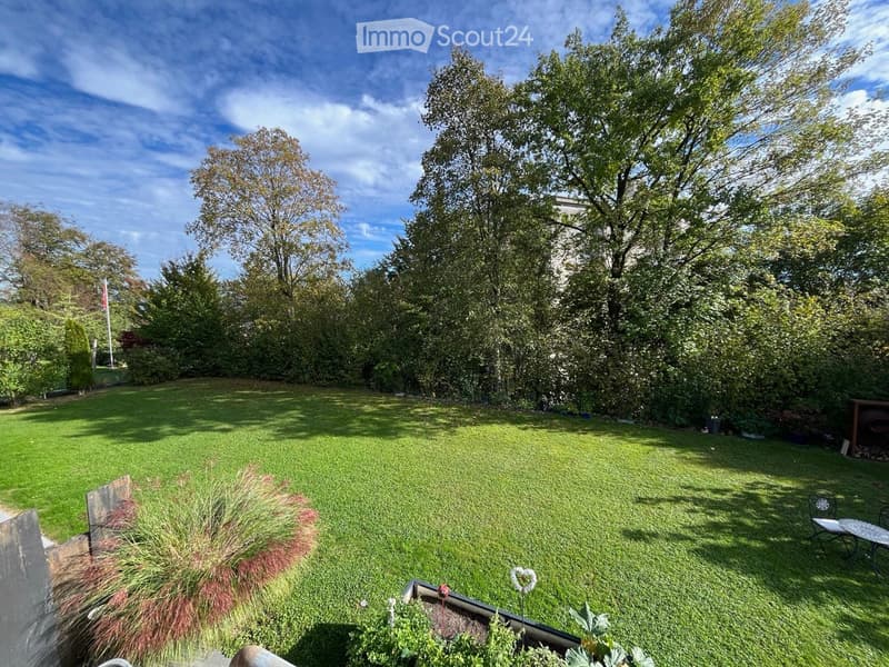 Well-maintained garden with a large lawn, trees, shrubs, and a small seating area with a table and chairs.