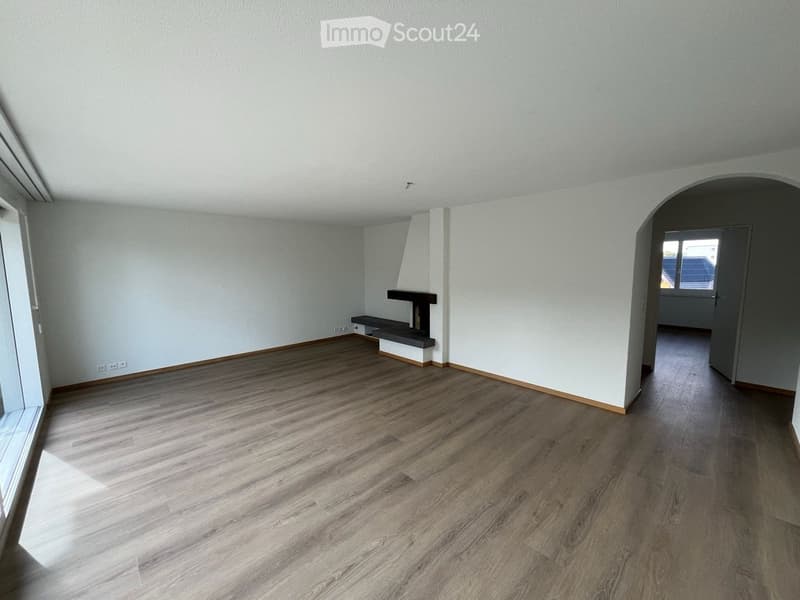 Empty room with light-colored wooden flooring, white walls, and a small built-in bench and fireplace. The room has large windows allowing natural light to enter.