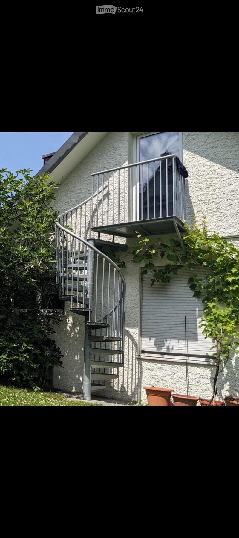 Spiral staircase leading to a balcony with a metal railing, surrounded by greenery and potted plants.