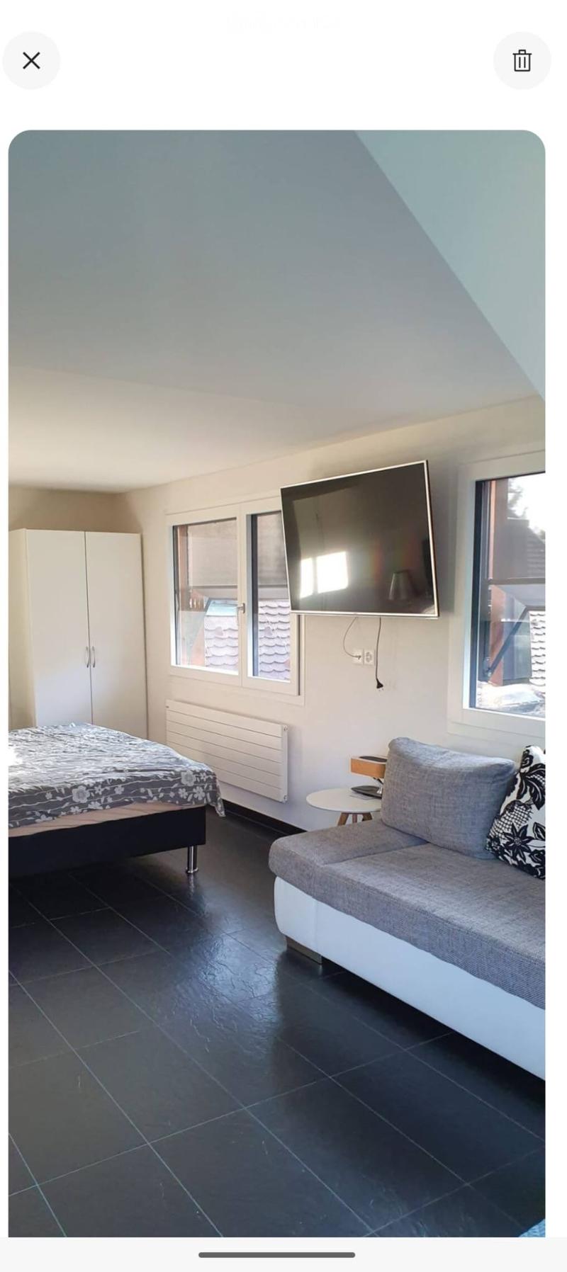 A modern, furnished room with a bed, sofa, small table, chair, television, and cabinet. The room features dark tiled flooring and large windows.