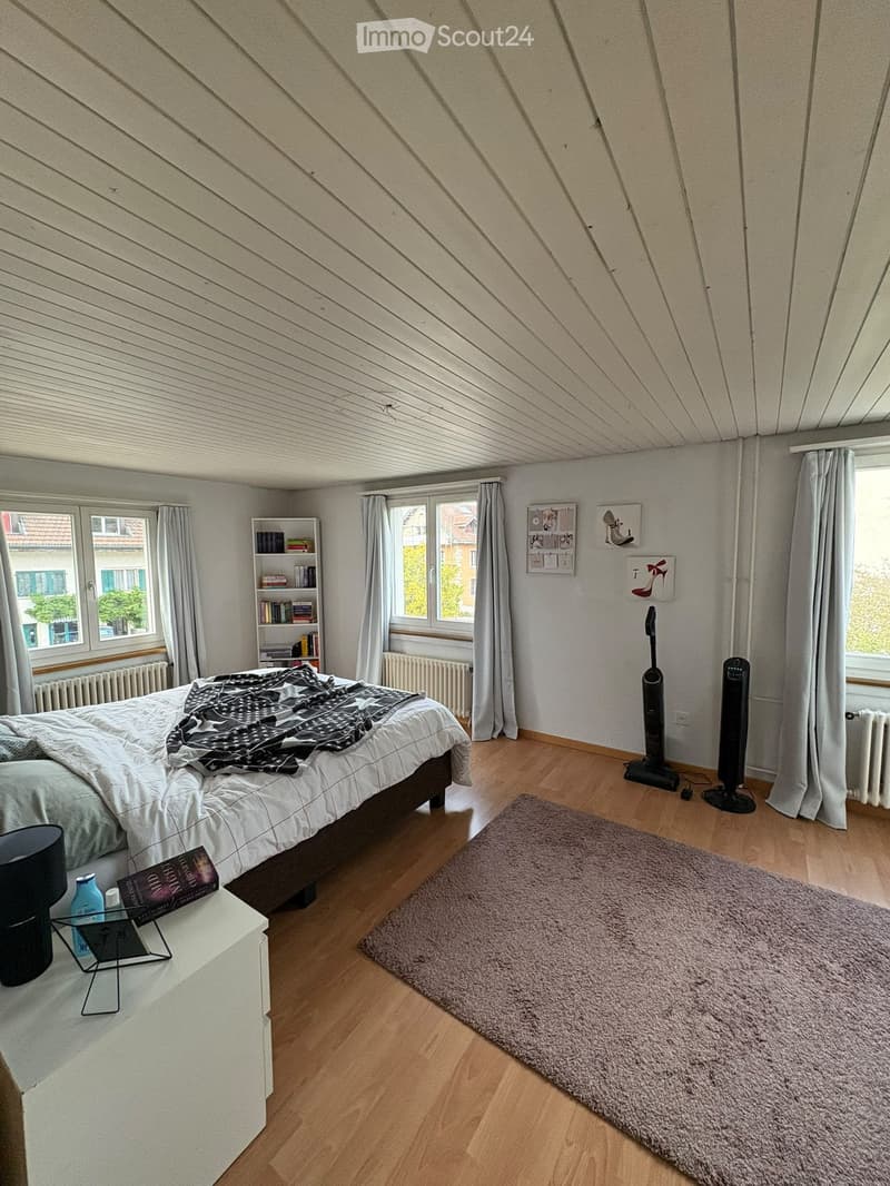 Bright bedroom with wooden flooring, slanted ceiling, large windows with curtains, bed with bedding, nightstand, bookshelf, and decorative items on walls.