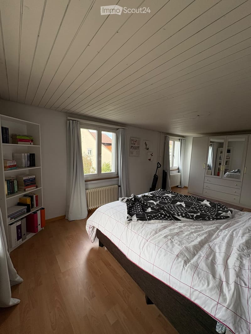 Bedroom with wooden floor, slanted wooden ceiling, large window with curtains, bed with white and black star-patterned blanket, bookshelf with books, mirror cabinet, radiator, and vacuum cleaner in corner.