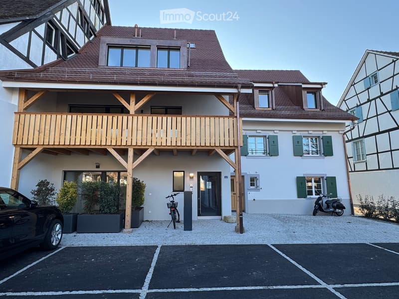 Two-story house with a wooden balcony, green shutters, and a parking area with a car and scooter.