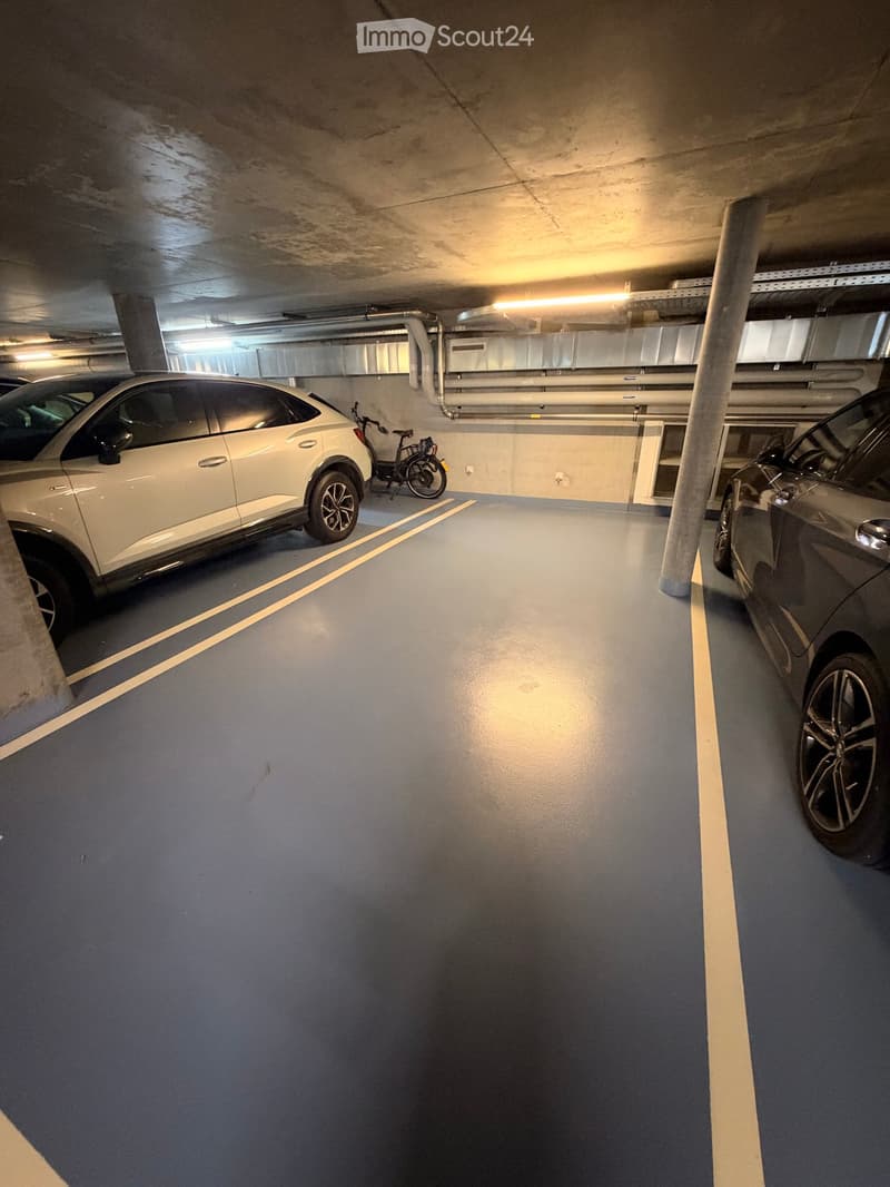 Underground parking area with marked parking spaces, a white car, a bicycle, and a dark-colored car partially visible.