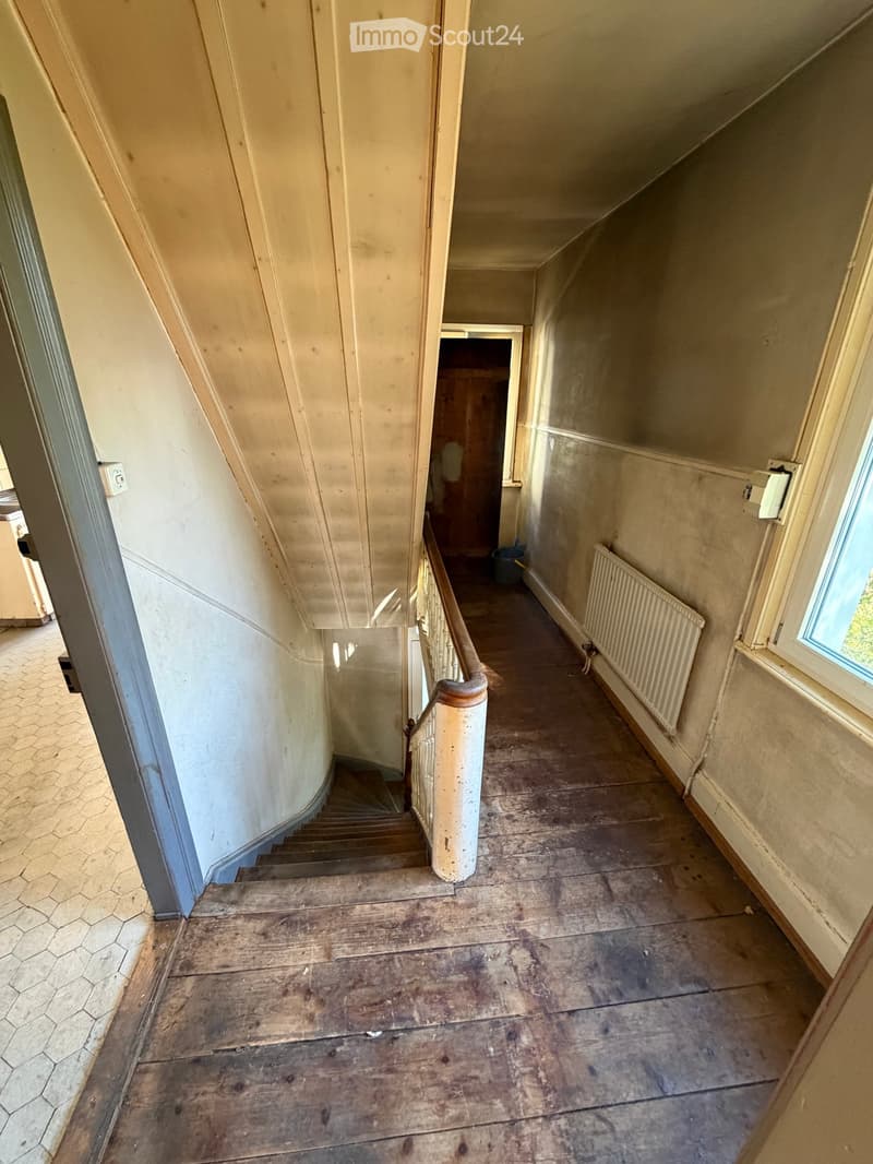 Empty hallway with wooden flooring, a staircase leading upwards, and a radiator on the right wall. The walls are painted in a light color, and there is a window at the end of the hallway.
