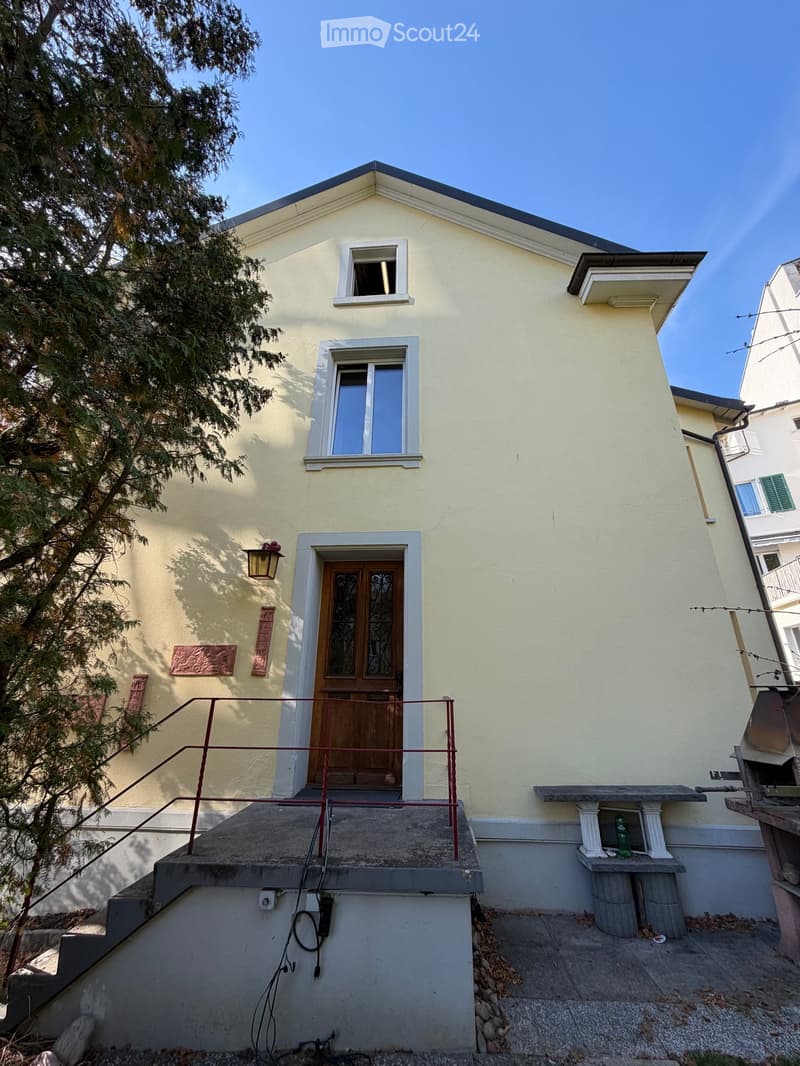 Two-story house with light yellow exterior, white-framed windows, wooden front door with decorative glass panels, small balcony with railing, and a stone bench on the side.