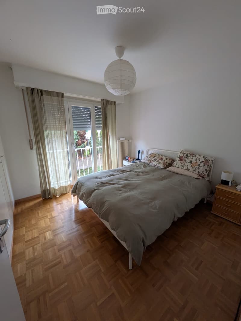 Cozy bedroom with wooden flooring, a large bed with patterned pillows and a fur throw, a nightstand with a lamp, and large windows with curtains leading to a balcony.