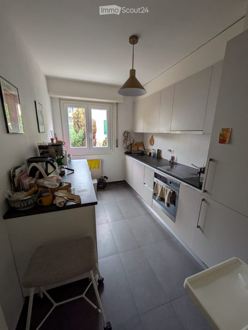 Modern kitchen with white cabinets, grey tiled floor, and a central island with a black countertop. Visible items include a stool, coffee maker, cups, and a framed picture on the wall.