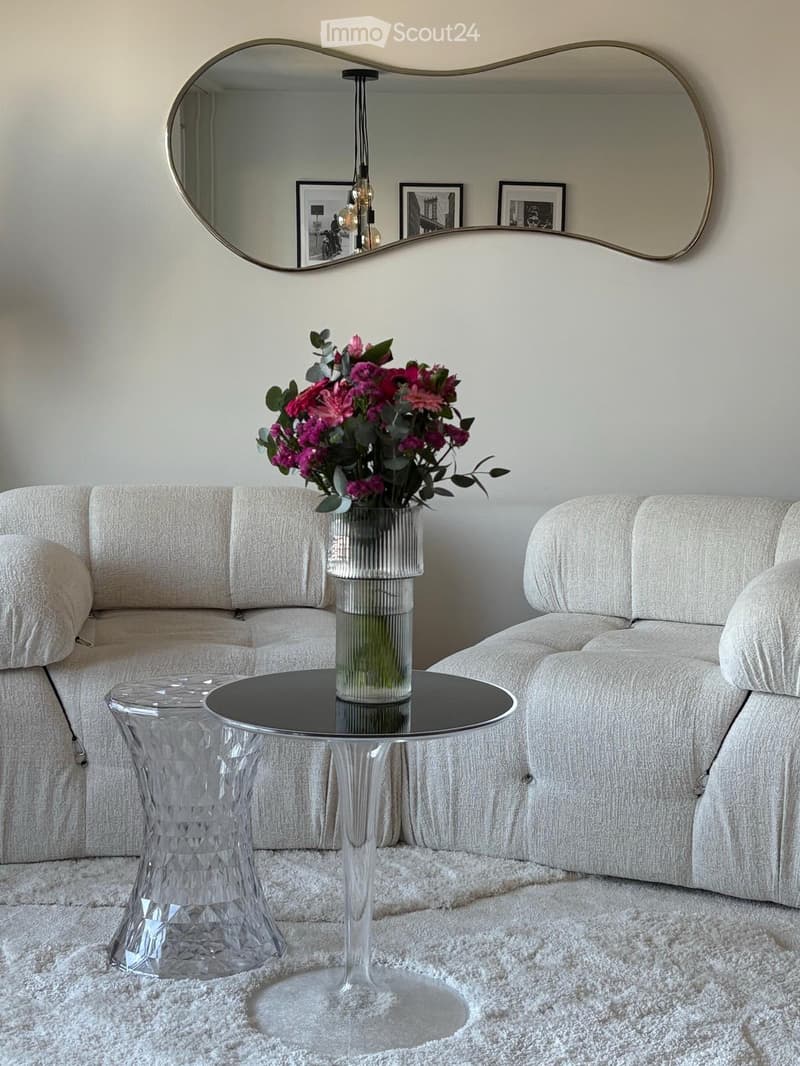 Living room with a light grey sofa, a round black coffee table with a vase of pink flowers, a clear glass side table, and a large decorative mirror on the wall.