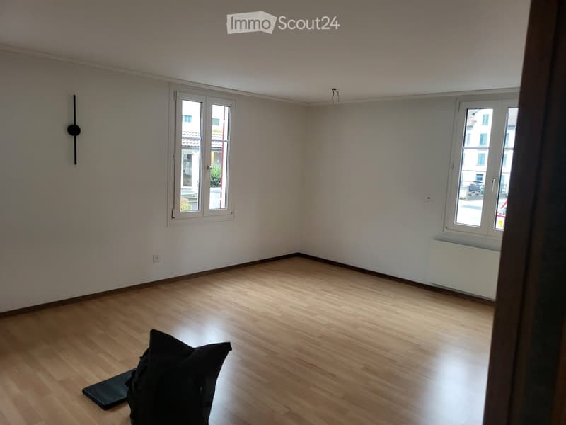 Empty room with wooden flooring, white walls, and two windows offering a view of the street.