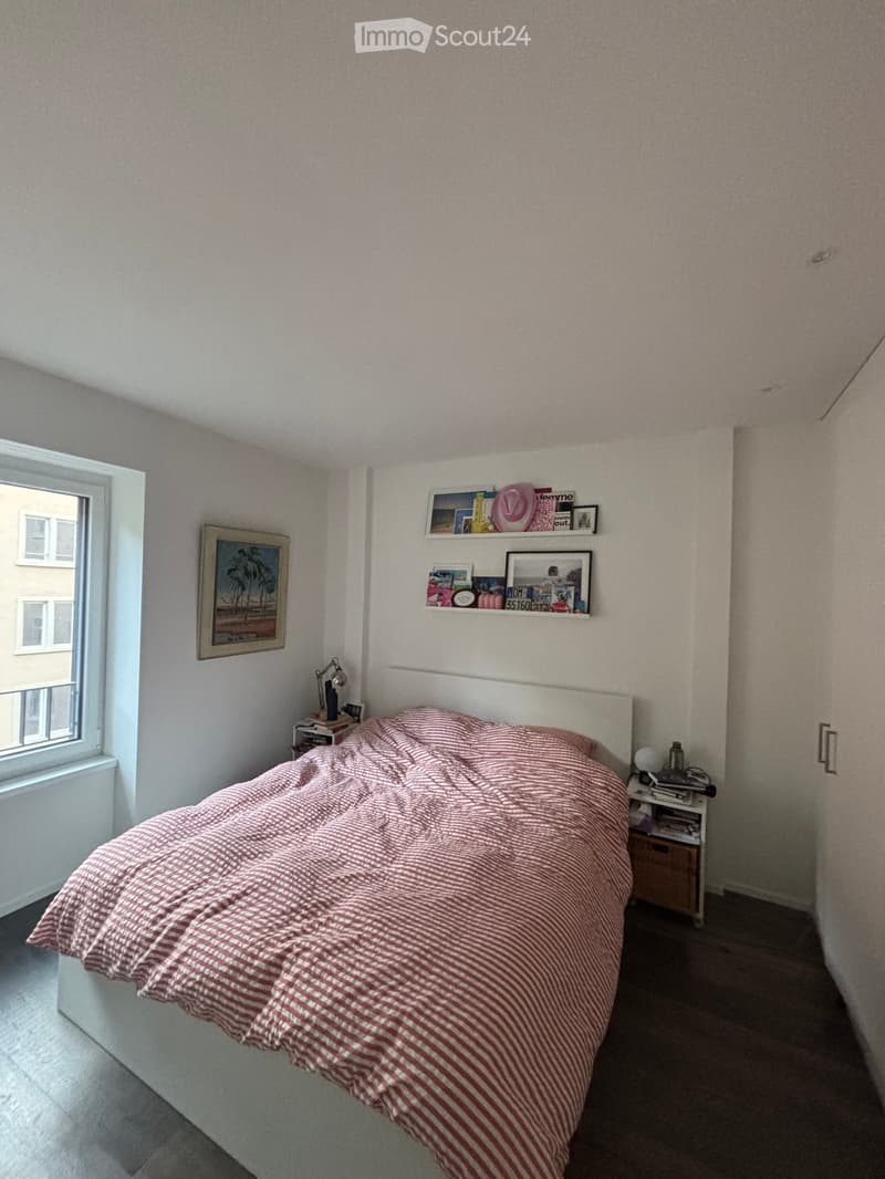 Cozy bedroom with a neatly made bed, nightstand, lamp, and wall-mounted shelf with picture frames and decorative items.