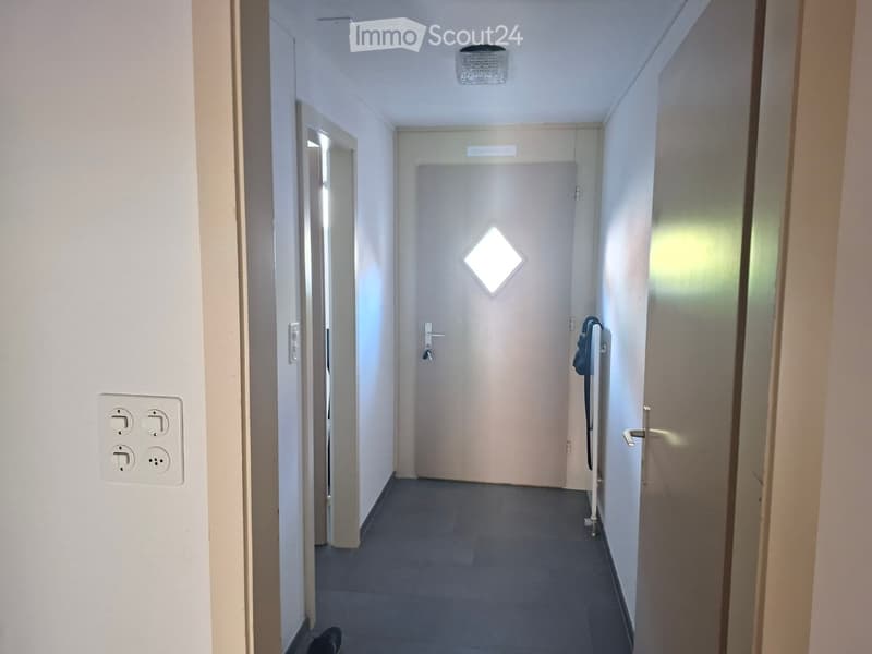 A narrow hallway with white walls, a closed door at the end, and a light fixture on the ceiling.