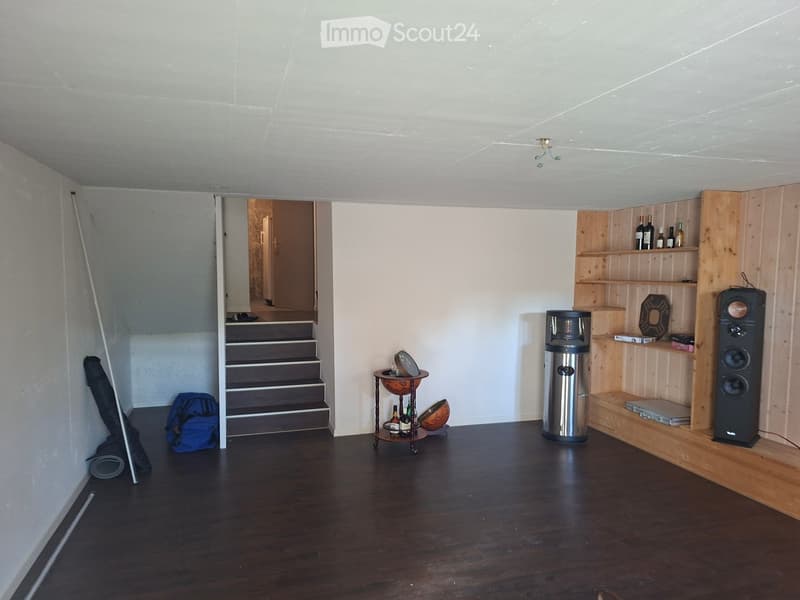 A modern, sparsely furnished room with wooden flooring, a staircase leading upwards, a wooden shelf with a speaker and decorative items, a small round table with bottles, and a backpack on the floor.