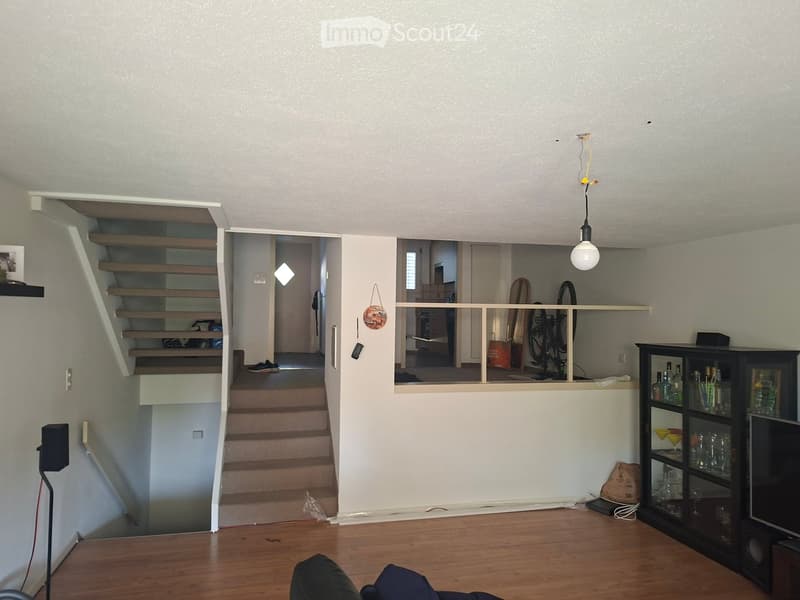 The image shows a modern living room with wooden flooring, a staircase leading to an upper level, and a balcony overlooking the living area. Visible objects include a cabinet with glass doors, a screen, a stool, and a bicycle in the background.