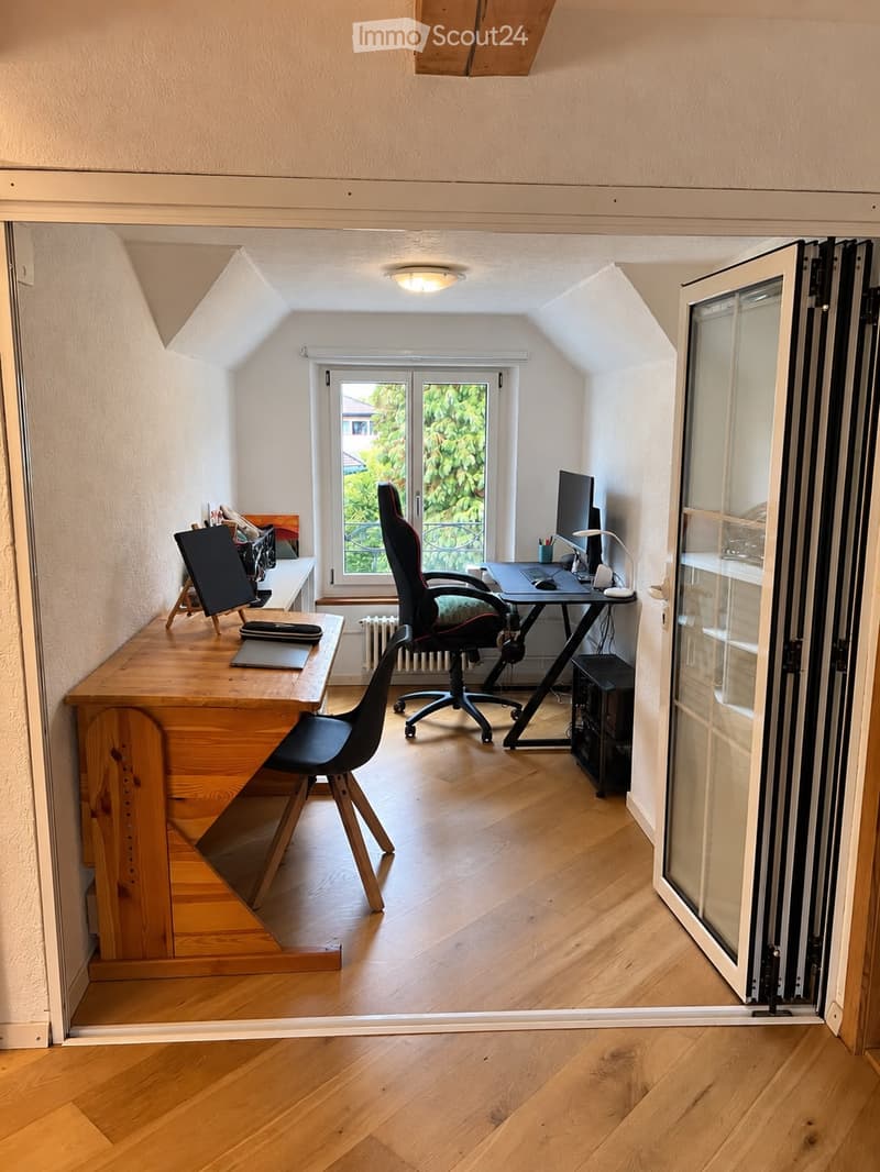 A well-lit, modern workspace with wooden flooring, a large window, a desk with a laptop, a comfortable chair, and a secondary table with a laptop and screen.
