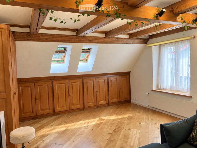 Bright attic room with wooden beams, exposed wooden ceiling, skylights, large window with white curtains, built-in wooden cabinets, and a small sofa.