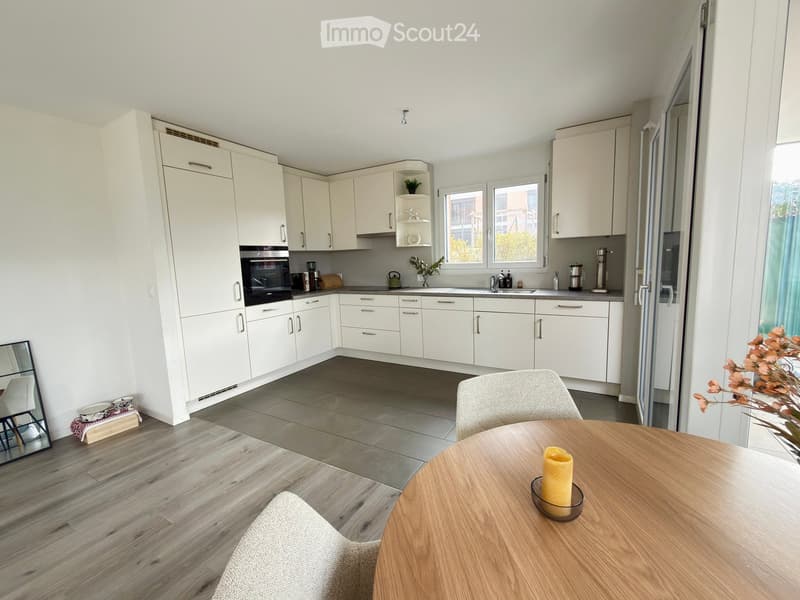 Modern kitchen with white cabinets, wooden table, and grey tiled floor. Includes a sink, oven, and a small dining area with a chair and candle.