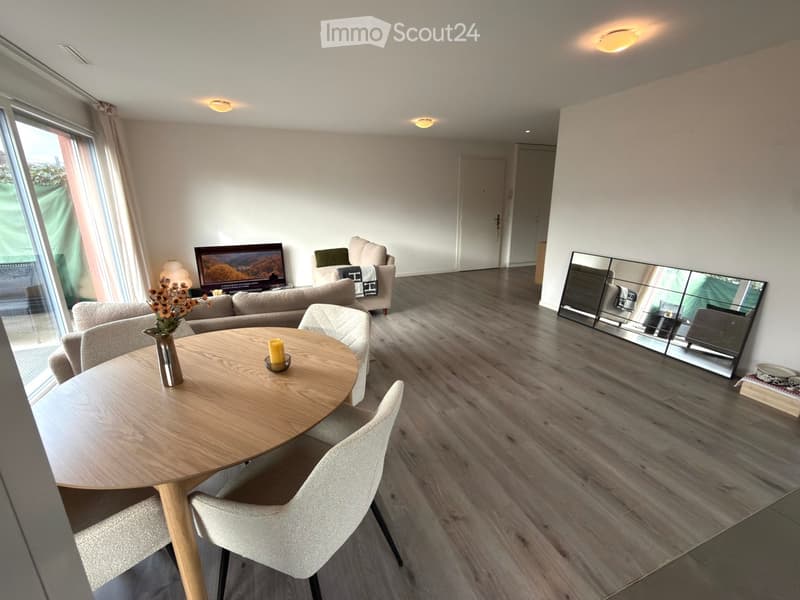 Modern living room with a round wooden table, sofa, chairs, mirror, and television. Features large windows with curtains and a vase with flowers on the table.