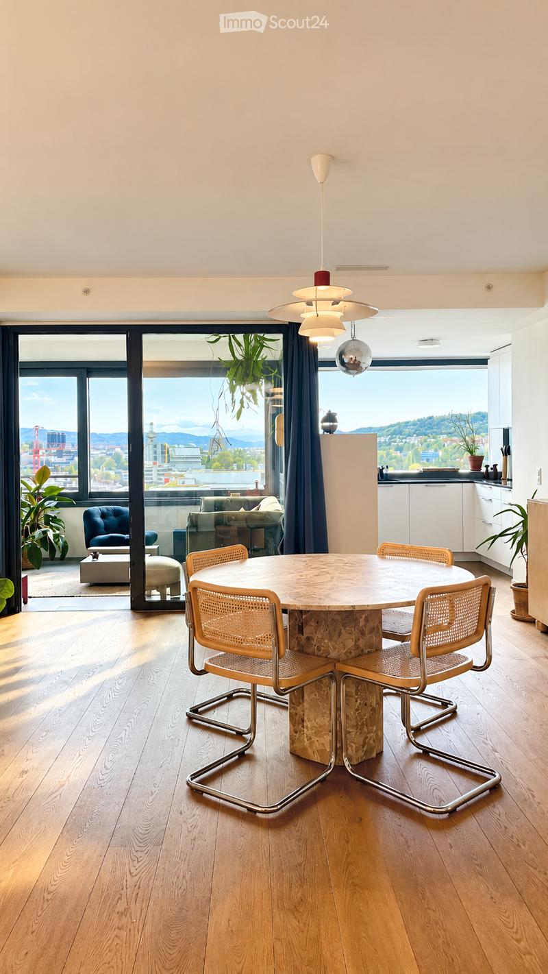 Modern dining area with a round marble table, four wicker chairs, pendant lighting, and a view of the cityscape through large windows. A small seating area with a sofa and ottoman is visible through the open doorway.