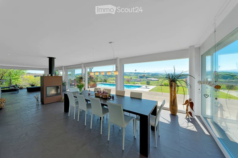 Modern dining area with a sleek black dining table and white chairs, a central fireplace, large windows offering a scenic view, and decorative vases and items on the table and floor.